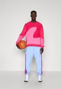 Rosa og rød sweatshirt med bølgedesign, parret med lyseblå sweatpants med lilla detaljer. Sort basketball holdt i venstre hånd.