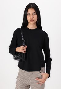 Svart langermet ribbestrikket topp med rund hals, kombinert med rutete bukser. Holder en liten svart lakk skulderveske. Glatt, tettsittende design.