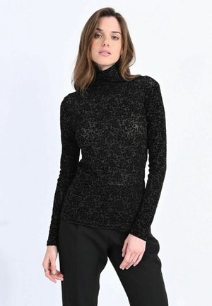 Donna con capelli castano chiaro che indossa una maglia nera a collo alto con motivo e maniche lunghe e pantaloni neri su uno sfondo chiaro uniforme.