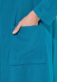 Garbamento in cotone teal con tasca frontale. Tessuto morbido con una finitura liscia e maniche arrotolate, evidenziando elementi di design pratico.