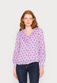 Blusa lilás clara com decote franzido, apresentando uma estampa de flores e geométrica em roxo escuro, e mangas franzidas. Estilizada com jeans escuros.