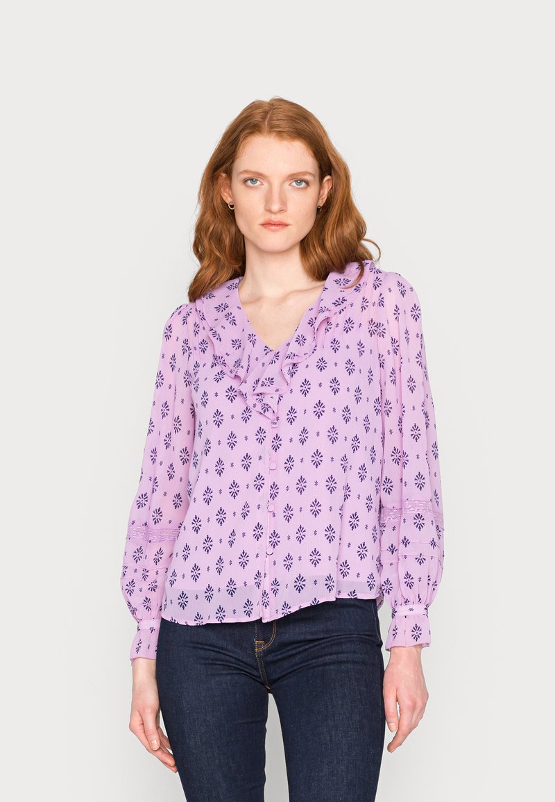 Blusa lilás clara com decote franzido, apresentando uma estampa de flores e geométrica em roxo escuro, e mangas franzidas. Estilizada com jeans escuros.