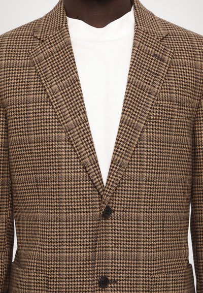 Brown houndstooth-patterned blazer with notch lapels, two black buttons, and a textured fabric. Worn over a white crew-neck shirt.