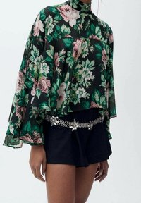 Blouse en voile à motifs floraux sur fond noir, avec des motifs de fleurs roses et vertes, accompagnée de shorts noirs et d'une ceinture en chaîne argentée ornée d'accents floraux.