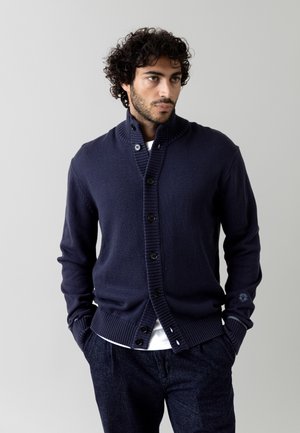 Marineblaue Strickjacke mit geripptem Kragen und Bündchen, fünfknöpfiger Vorderseite und strukturiertem Stoff; über einem weißen Hemd getragen, kombiniert mit dunklen Hosen.
