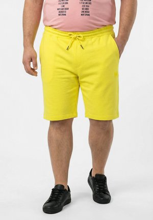 Man draagt felgele shorts met trekkoord, een roze T-shirt met zwarte tekst en zwarte sneakers, staand met één hand in de zak.