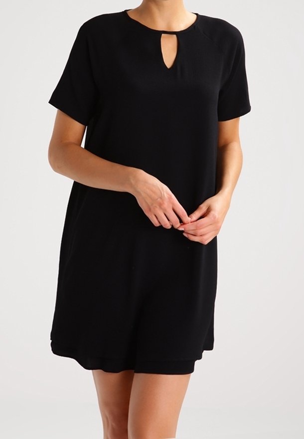 Femme portant une robe noire à manches courtes avec un décolleté goutte d'eau, debout les mains doucement jointes devant un fond blanc uni.