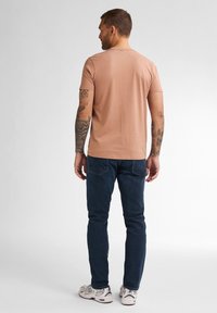 Tričko s krátkým rukávem v světle růžové barvě s kulatým výstřihem, v kombinaci s tmavě modrými džínami a bílými teniskami. Viditelné tetování na paži.