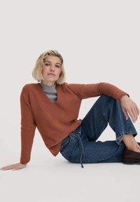 Ein rötlich-brauner, gerippter Strickpullover mit V-Ausschnitt, kombiniert mit lockeren, ausgewaschenen blauen Jeans. Das Modell sitzt und zeigt den entspannten Schnitt und die Textur.