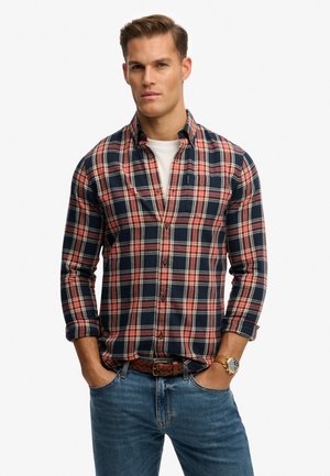 Homme portant une chemise à carreaux bleu marine, rouge et blanche boutonnée, sur un t-shirt blanc, jean bleu, ceinture tressée marron et montre-bracelet en or, les mains dans les poches.