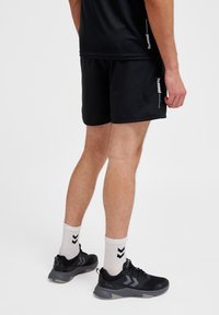Svarta träningsshorts med en slät yta, i kombination med svarta sneakers och vita strumpor med ett chevronmönster, sedd bakifrån.