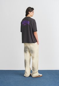 Løs sittende svart t-skjorte med lilla "Reebok"-logo på ryggen, kombinert med kremfargede bukser med en gradient grå design.