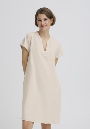 Femeie într-o rochie lejeră bej cu mâneci scurte și decolteu în V, stând cu mâinile la spate, zâmbind pe un fundal simplu, deschis la culoare.