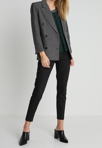 Blazer à double boutonnage en vichy noir et blanc, associé à un haut vert foncé et un pantalon noir slim, complété par des mules noires à talons carrés.