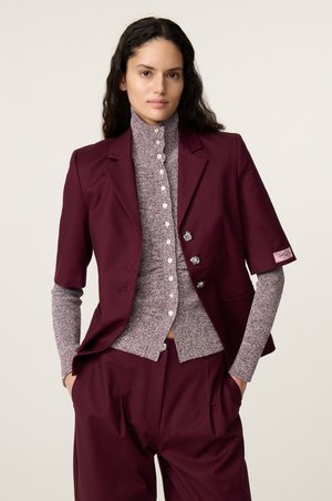 Vrouw met lang donker haar die een paarse korte mouwen blazer draagt over een geknoopt, getextureerd hoognekkig shirt en bijpassende paarse broek, handen in de zakken.