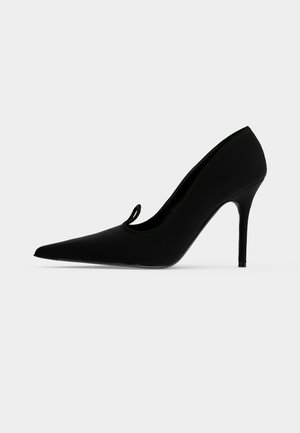 Sort spids hæl med stiletthæl og lille løkke detalje på oversiden, vist fra venstre profil.