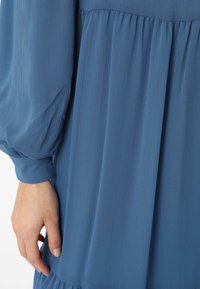 Blue, textured dress with gathered detailing and long, puffed sleeves. Fabric appears lightweight and flowing, showcasing a soft sheen.