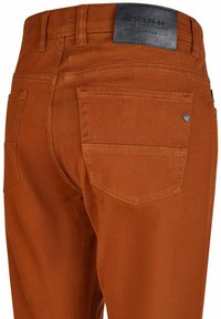 Rdzawoczerwone spodnie o gładkiej fakturze, z dwiema tylnymi kieszeniami i skórzaną naszywką z logo. Widoczne szycie maszynowe wzdłuż szwów.