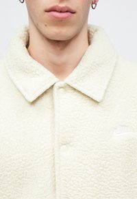 Λευκό μπουφάν από φλις με γιακά, κλείσιμο με κουμπιά και κεντημένο λογότυπο στο αριστερό στήθος. Απαλή υφή με ελαφριά γυαλάδα.