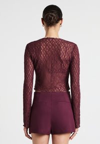 Burgunderfarbene lange Spitzenbluse mit einem cropped Design, die ein florales Muster und einen offenen Rücken aufweist, kombiniert mit hochgeschnittenen Shorts.