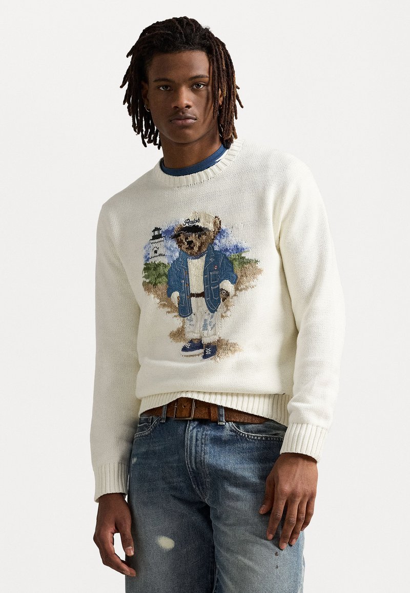 Krém kötött pulóver, amelyen egy kék kabátot és sapkát viselő medvét hímeztek, szakadt kék farmernadrággal és barna övvel párosítva.