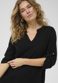 Czarny lekki top z dekoltem w serek, z podwiniętymi rękawami przymocowanymi guzikami i miękką fakturą. Modelka pozuje lekko z boku.