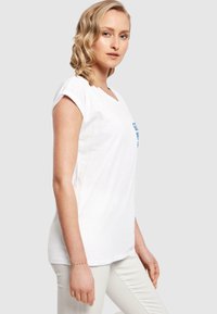 Weißes Baumwoll-T-Shirt mit V-Ausschnitt und aufgerollten Ärmeln. Mit blauer Schrift auf der Vorderseite. Einfaches Design und weiche Textur. Kombiniert mit hellen Hosen.