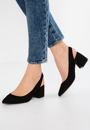 Person iført blå jeans og sorte ruskindsslingback hæle med blokhæl på hvid baggrund.