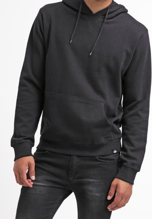Mann trägt einen schlichten schwarzen Hoodie mit Fronttasche und Kordelzügen, kombiniert mit schwarz gewaschenen Jeans.