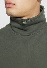 Gros plan d'une personne portant un pull à col montant vert olive avec un cordon de serrage réglable et un toggle au niveau du col.