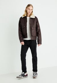 Brown leather jacket with a white shearling collar, front pockets, a zip closure, paired with black jeans and grey athletic shoes.