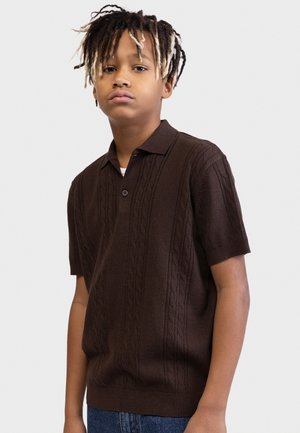 Jaunas žmogus trumpais dredekais, dėvintis tamsiai rudą trumpomis rankovėmis megztą polo marškinėlį ir mėlynas džinsines kelnes, žiūrintis tiesiai į priekį su neutraliu veido išraiška.