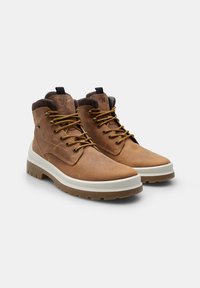 Botas de cuero marrón hasta el tobillo con suelas beige, punteras redondas y cordones marrones. Presentan un cuello acolchado y un logo grabado en el lateral.