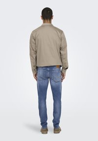 Φωτεινό μπεζ μπουφάν με γιακά, συνδυασμένο με μπλε τζιν που διαθέτουν πίσω τσέπες και χρυσή ραφή. Το μοντέλο φαίνεται από την πλάτη.