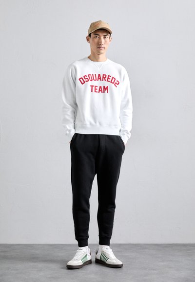 Man wearing beige cap, white sweatshirt with red "DSQUARED2 TEAM" text, black pants, and white sneakers with green stripes standing against a plain wall.