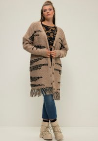 Beige Strickjacke mit schwarzen Streifen und Fransen- Saum, mit Knöpfen vorne. Getragen über einem gemusterten Oberteil, kombiniert mit blauen Jeans und Stiefeln.