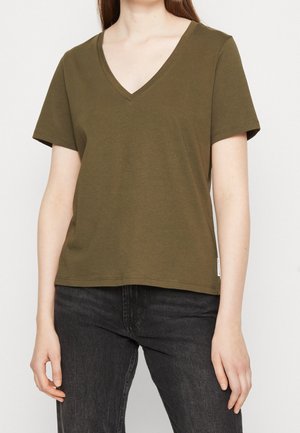 Kvinde iført olivengrøn kortærmet T-shirt med V-hals og sorte jeans, stående mod en ensfarvet baggrund.