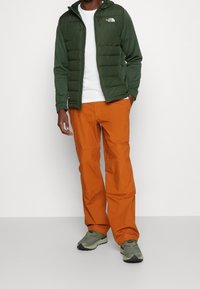 Vihreä puffatakki valkoisen t-paidan päällä, yhdistettynä oransseihin housuihin ja harmaisiin lenkkareihin. Takissa on huppu ja vetoketjulliset taskut.