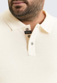Crèmekleurig polo shirt van textuur, ribbelstof. Voorzien van een messing met twee knopen en een zijlabel met merkinformatie.