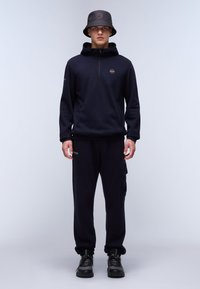 Felpa in pile blu marina con mezza zip, abbinata a pantaloni jogger coordinati con una tasca cargo. Accentuata con un cappello a secchiello nero. Stivali neri.