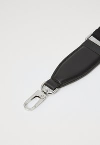 Pulseira de couro preto com fecho de metal prateado, presa a uma banda de tecido sobre um fundo branco.