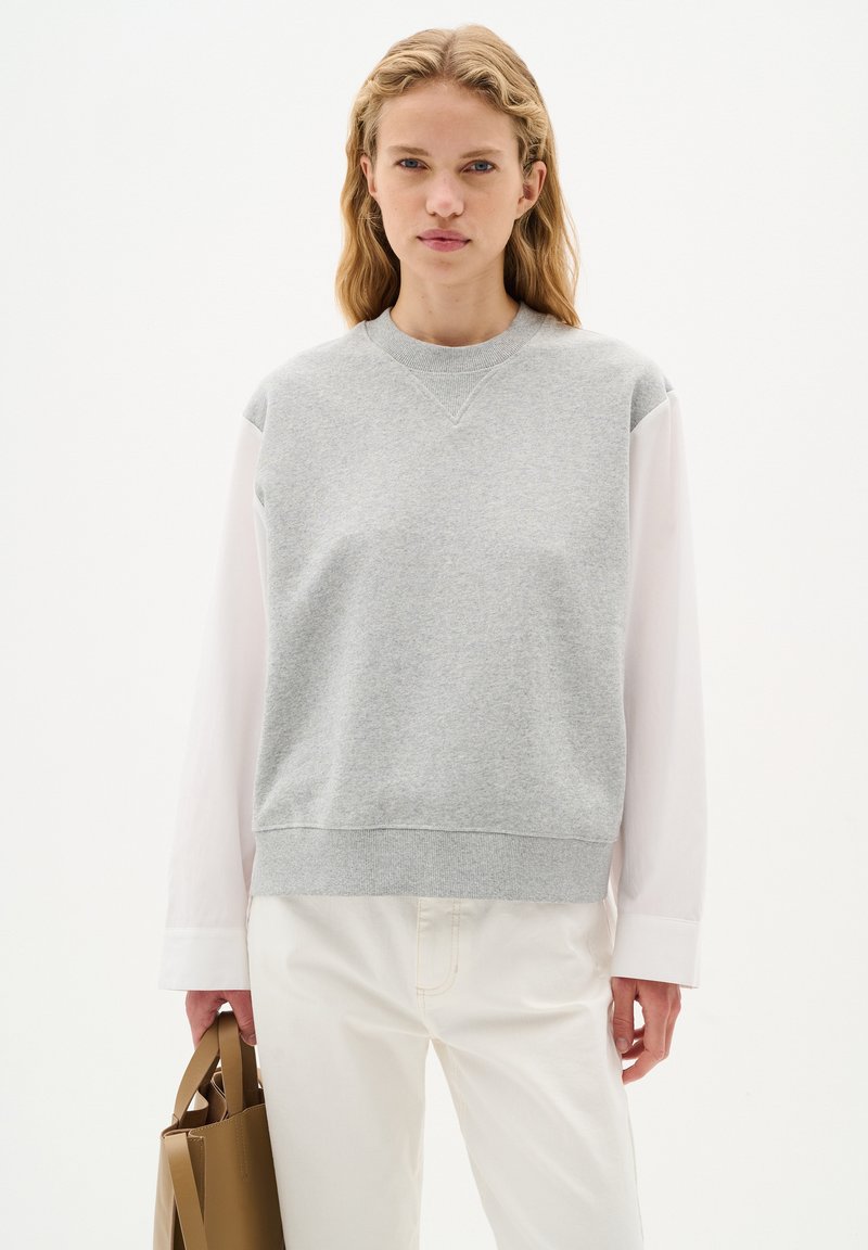 Lichtgrijze sweatshirt met een ronde hals, lange witte mouwen, ribgebreide manchetten en onderkant, gecombineerd met witte broek en een bruine tote bag.