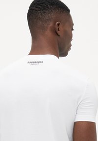 Valkoinen lyhythihainen t-paita, joka on valmistettu sileästä puuvillasta. T-paidassa on musta logo takana kauluksessa. Suunnittelu on tyylikäs ja minimalistinen.