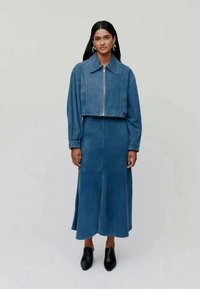 Veste en denim à manches courtes avec une fermeture éclair à l'avant, associée à une longue jupe bleue unie. Le modèle porte des chaussures à talons noires. Texture lisse, coupe ajustée.
