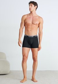 Zwarte katoenen boxershorts met een gladde textuur, met een Calvin Klein-logo op de tailleband, model staat op een neutraalgekleurde vloer.