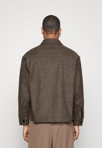 Braune Wolljacke mit spitzem Kragen, abgesenkten Schultern und geknöpften Manschetten. Die Rückseite ist schlicht und minimalistisch gestaltet.
