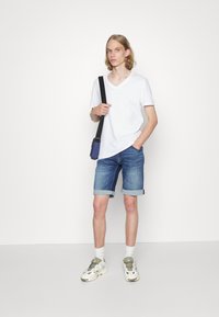 T-shirt bianco a scollo a V, pantaloni corti in denim blu con orli arrotolati, sneakers beige e grigie, e una piccola borsa a tracolla blu.