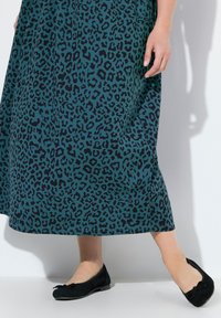Robe teal avec imprimé léopard noir, en tissu fluide et longueur cheville ; associée à des ballerines en daim noir avec un petit nœud.