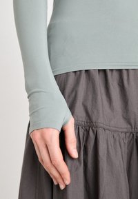Long-sleeve, light green top with fitted texture and thumbhole cuffs, paired with a gray, gathered skirt featuring a smooth waistband.
