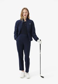 Veste rembourrée bleu marine avec fermeture éclair, associée à un pantalon bleu marine sur mesure. Le modèle tient un club de golf, portant des chaussures et des gants blancs.